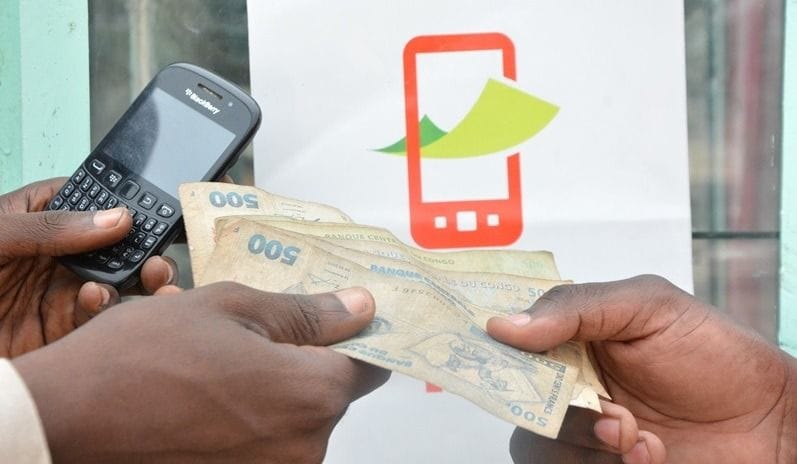 Deux personnes échangent des billets de 500 francs congolais, l’une tenant un téléphone portable, devant une affiche de service de mobile money.