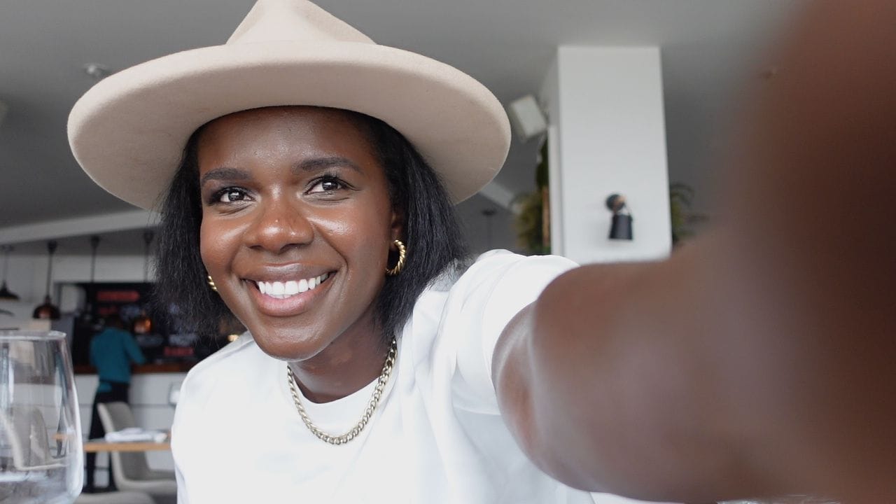 A smiling woman in a wide-brimmed beige hat and white T-shirt takes a selfie inside a bright café.